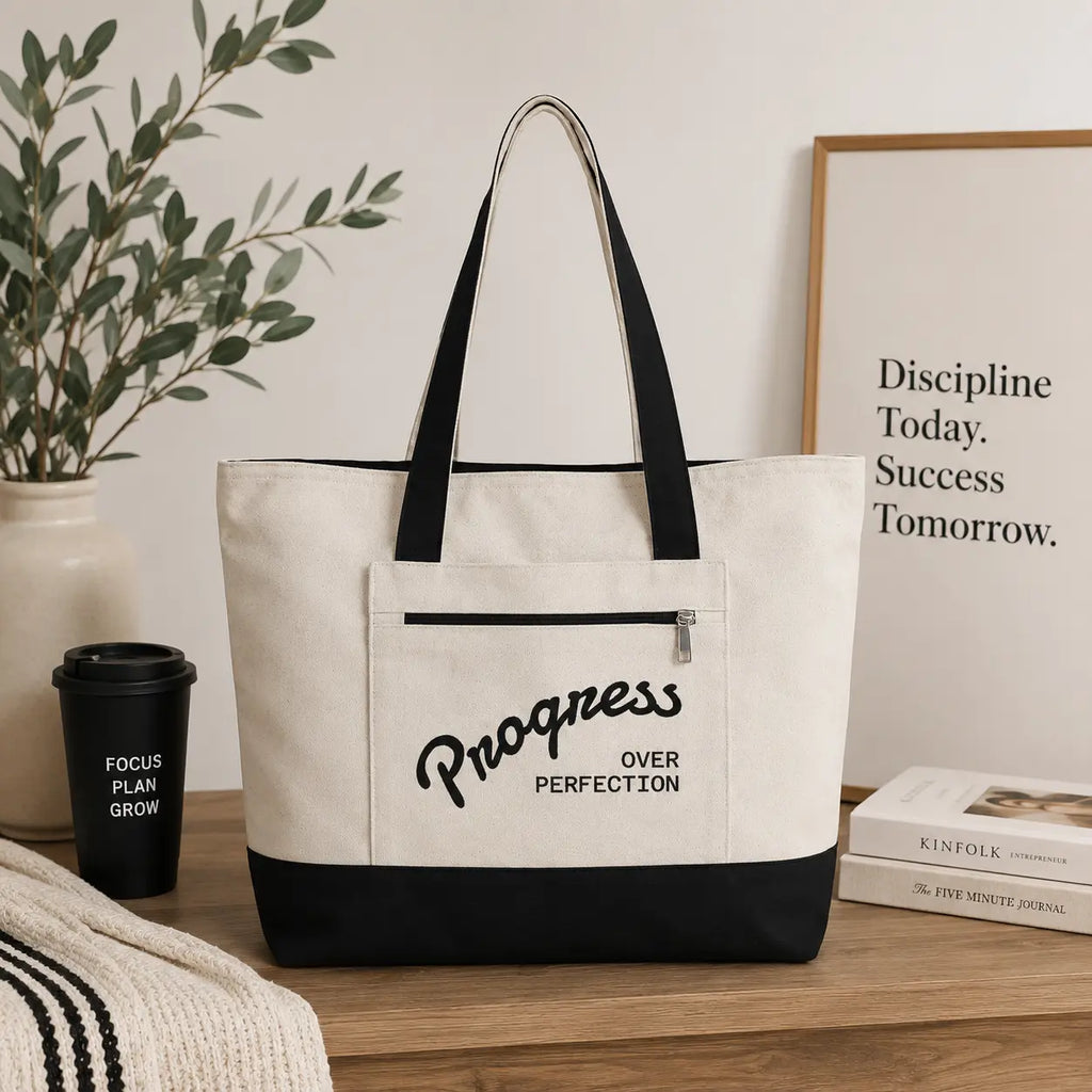 Progress Over Perfection Tote Bag | Minimalist canvas tote bag with black straps and zipper pocket featuring the motivational quote “Progress Over Perfection” in bold black typography on a neutral cream background.